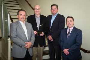 A group of four professionally dressed men, one holding an award, posing on a staircase