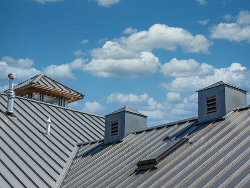 A light-gray metal roof with ventilation pipes and dormer windows set against a bright blue sky